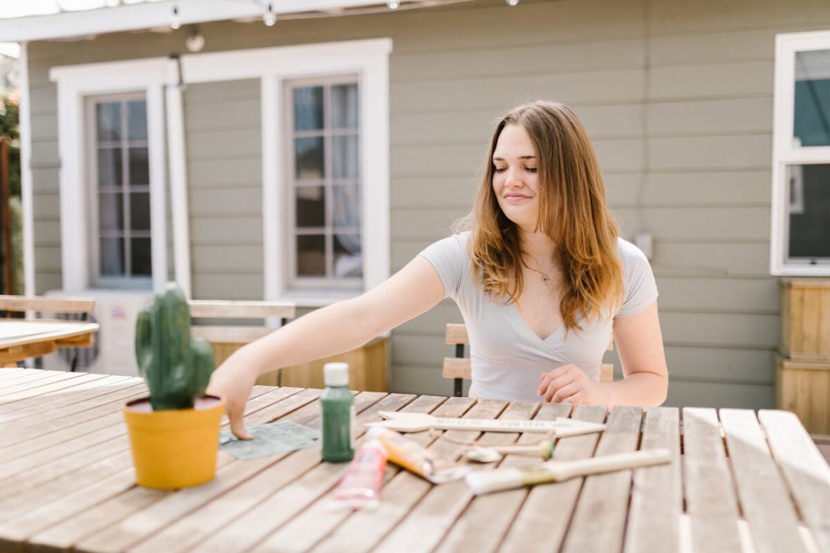 Young woman crafting outdoors with paints and brushes on a wooden table.